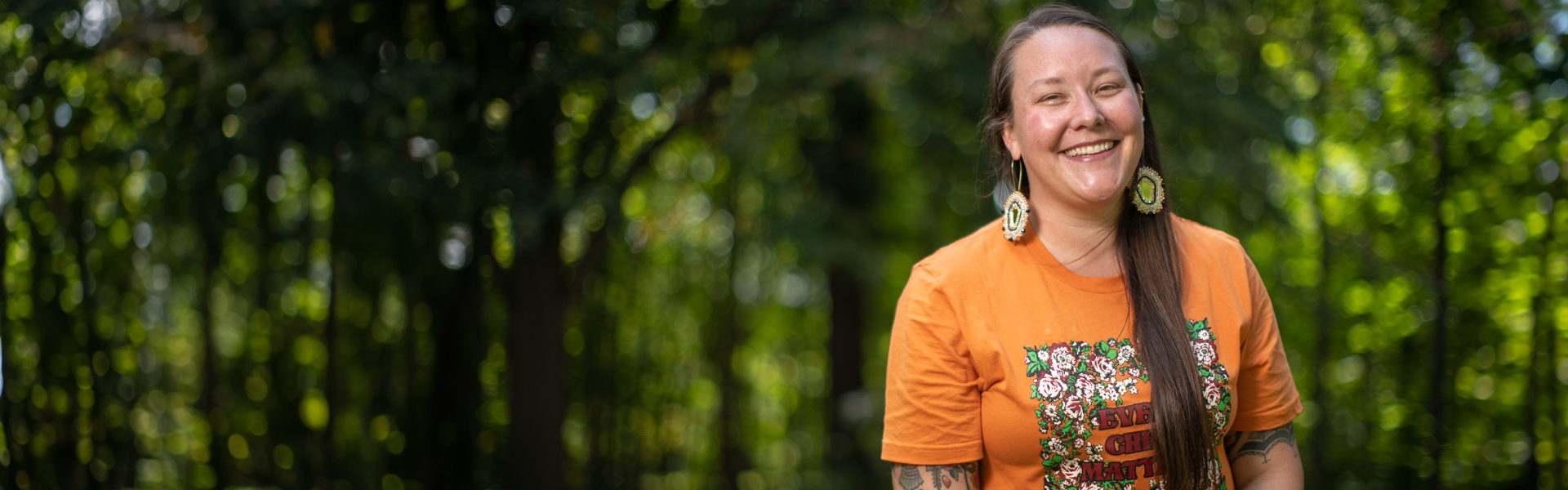 Photo of Indigenous woman smiling in front of greenery wearing an orange t-shirt